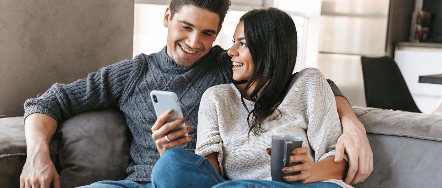 Paar, das auf dem Sofa sitzt und gemeinsam auf ein Smartphone blickt.