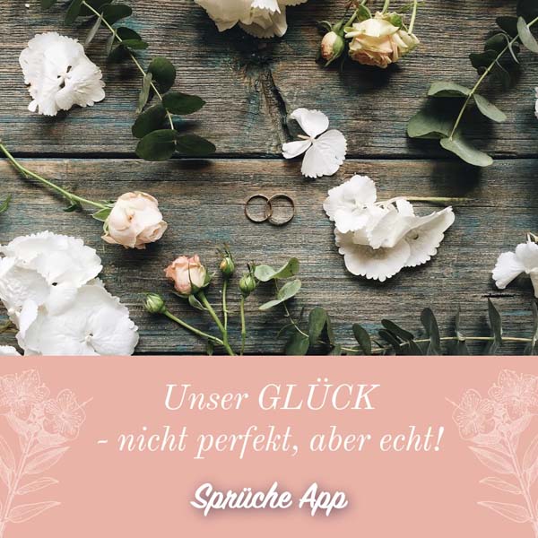 Eheringe umgeben von weißen Blüten, Rosen und Eukalyptus auf einer Holzoberfläche mit dem Spruch: „Unser Glück – nicht perfekt, aber echt!"